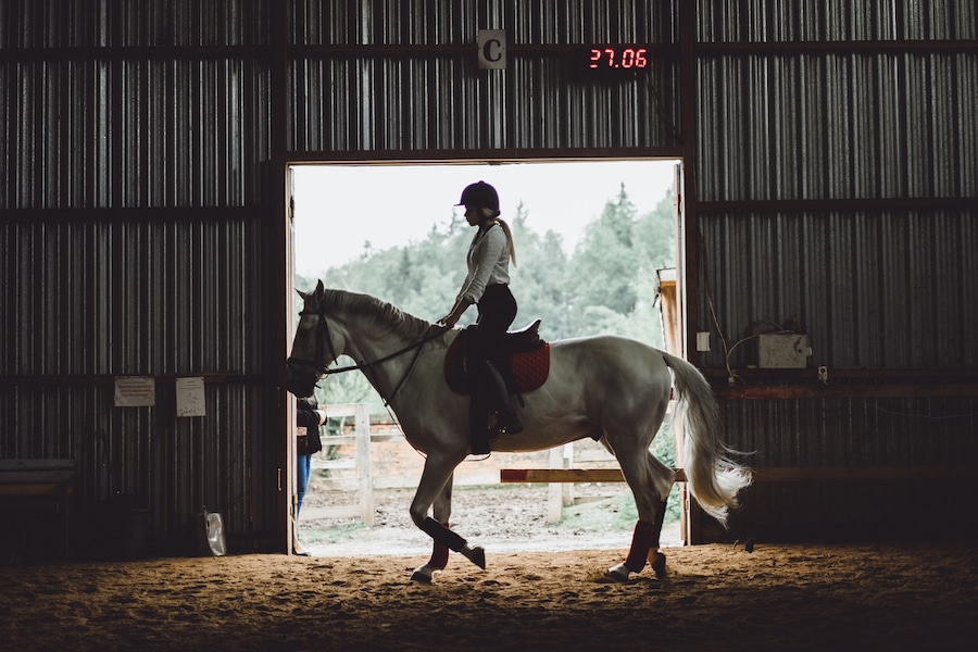 A woman riding a white horse in a barn with low dust arena footing.