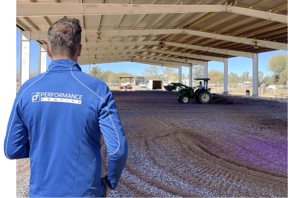 A man in a blue jacket standing next to a tractor in a horse arena. A man in a blue jacket standing next to a tractor in a horse arena.