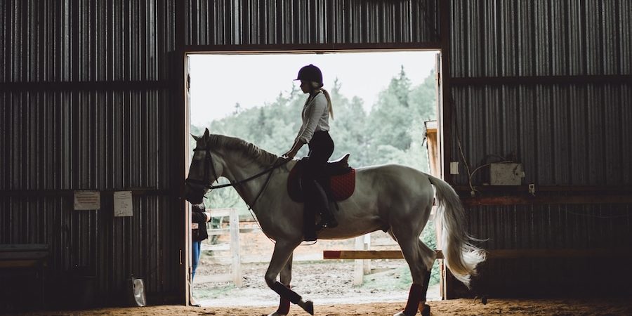A woman riding a white horse in a barn with low dust arena footing.